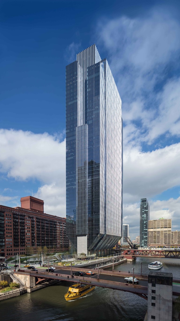 The full height of the tower seen from across the river, the base tapering to its footing between the tracks and the water.