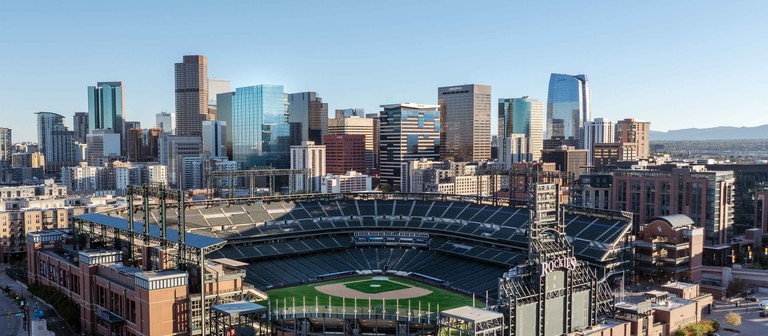 Looking out over Coors Field from above, with the downtown Denver skyline beyond and the mountains faint on the horizon.