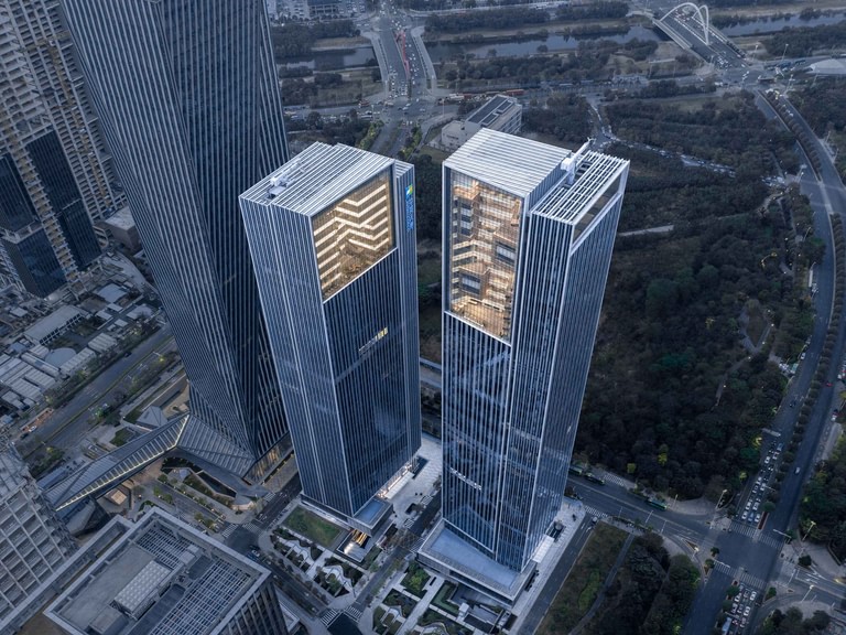 Aerial view of both towers from above, the sky terraces open at the crowns and the green belt along the eastern edge of the site.