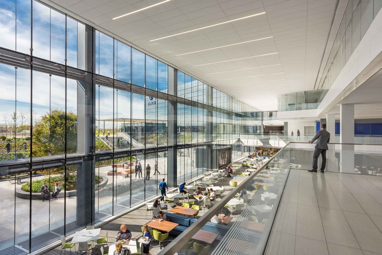The dining hall along a full-height glass wall, tables occupied and the plaza and landscape visible outside.