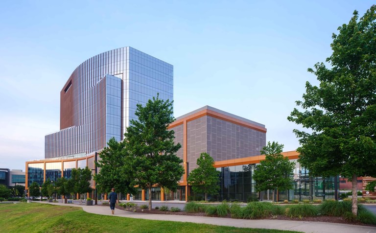 The building from the park side, glass storefronts and a low volume under a copper band with the tower behind.