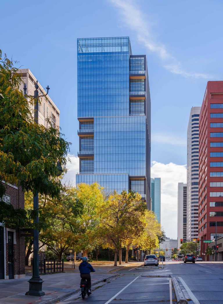 The tower seen from a LoDo street corner in autumn, brick low-rise neighbors and street trees in the foreground and a cyclist in the protected lane.