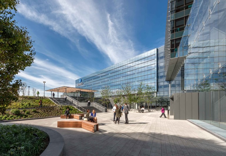 The plaza at grade, people seated on wood benches among planting with the glass facade and cantilever above.