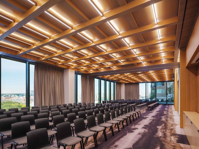 The conference center set up for a lecture, rows of chairs under a coffered wood ceiling threaded with light and a city view through the drapes.