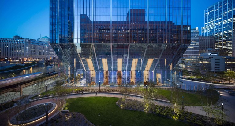 The underside of the building at dusk, the diagonal structural transfers lit behind the glass-fin wall above the park lawn.