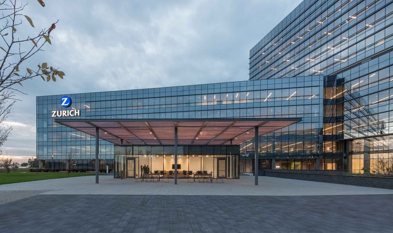 The main entry canopy at dusk, Zurich signage on the bar above and reception lit behind full-height glass.