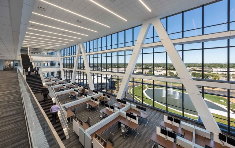 An office floor at the perimeter, workstations along the glass with the white diagonal bracing crossing it and the pond and prairie beyond.