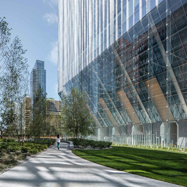 At grade alongside the glass-fin wall, diagonal members and spider fittings overhead, with a path and lawn running past.