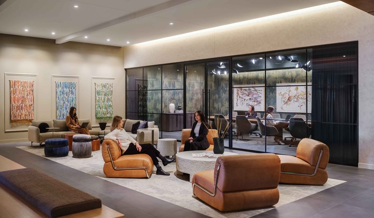 A lounge with tan leather seating and artwork on the walls, a glass-partitioned meeting room visible beyond.