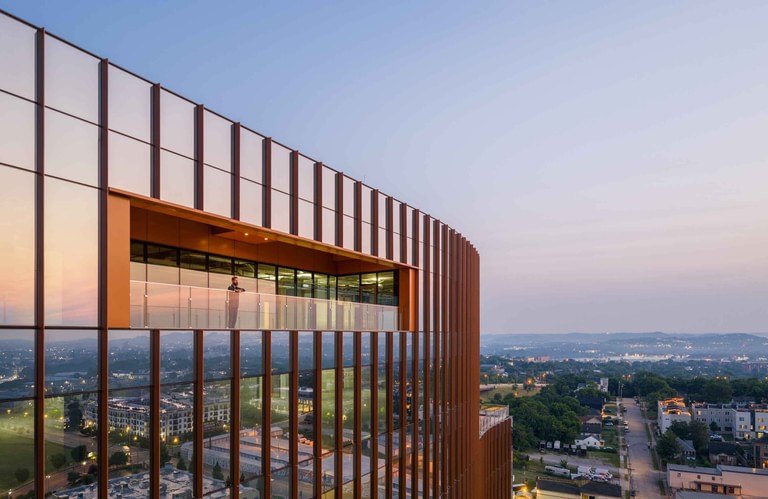 A terrace cut into the upper facade at dusk, lined in copper, with one figure at the glass rail and the city beyond.
