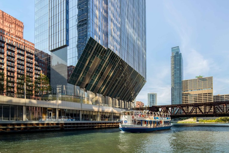 From the water, the angled underside lifting over the riverwalk with the restaurant glazing below and a tour boat passing.