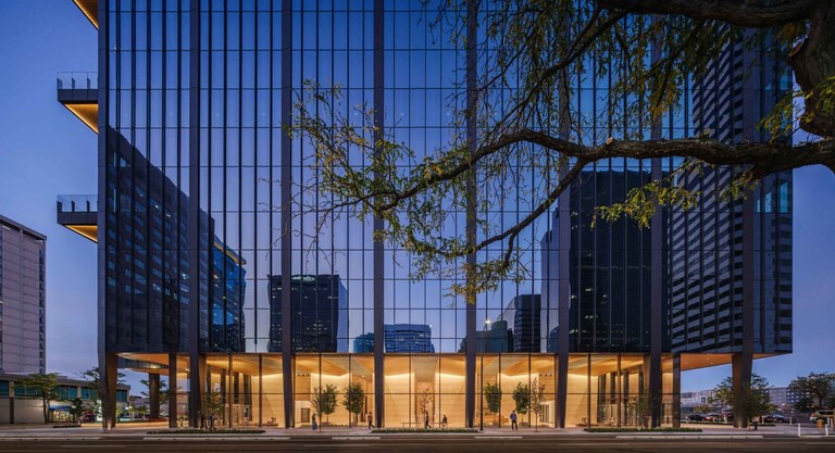The base of the tower at dusk seen through street trees, neighboring towers reflected in the glass and the lobby and retail lit at grade.