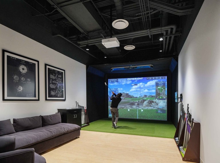 The golf simulator room, a player mid-swing at the screen with clubs racked along the wall and a sofa behind.