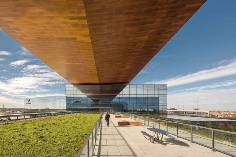 From the green roof of a lower bar, looking along the warm wood soffit of the cantilever overhead with a walking trail and railing at the edge.
