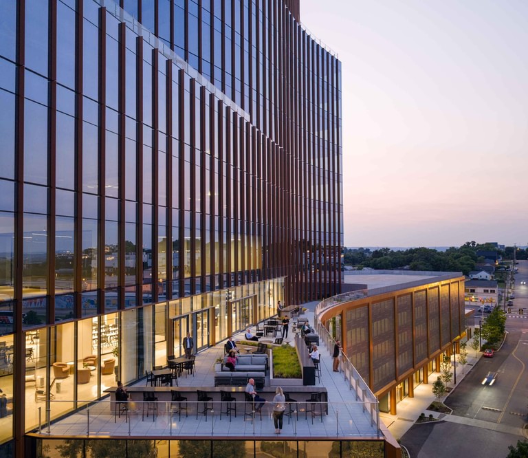 The amenity terrace in use at dusk, tables and planting following the curve of the facade with streets below.