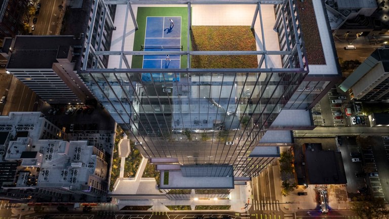 The rooftop activity deck from directly overhead, a blue pickleball court and green turf set against the city far below.